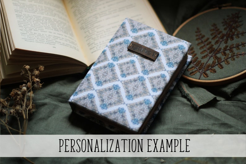 Puede incluir: Un libro cerrado con una cubierta estampada con un dise&ntilde;o geom&eacute;trico en tonos grises y azules. Una peque&ntilde;a etiqueta de madera con la palabra "TRAVEL" est&aacute; adjunta. El texto "PERSONALIZATION EXAMPLE" se muestra en la parte inferior de la imagen.