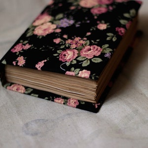 Puede incluir: Un libro de tapa dura con un dise&ntilde;o floral. La cubierta es negra con flores rosas, moradas y verdes. Las p&aacute;ginas est&aacute;n envejecidas y el libro descansa sobre una tela blanca.