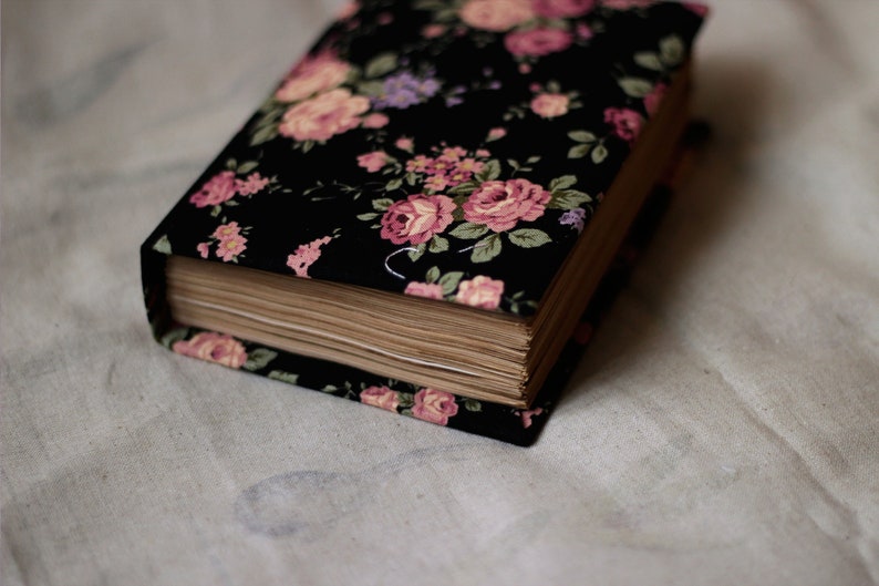 Puede incluir: Un libro de tapa dura con un dise&ntilde;o floral. La cubierta es negra con flores rosas, moradas y verdes. Las p&aacute;ginas est&aacute;n envejecidas y el libro descansa sobre una tela blanca.