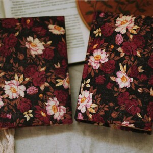 May include: Two black fabric journals with a floral pattern featuring pink, white and burgundy flowers. The journals are lying on a light brown surface.