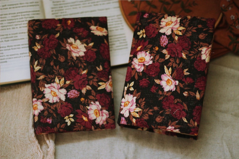 May include: Two black fabric journals with a floral pattern featuring pink, white and burgundy flowers. The journals are lying on a light brown surface.