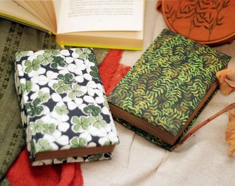 May include: Two hardcover journals with different patterned covers. One journal has a green and white clover pattern, and the other has a green and brown leafy pattern.
