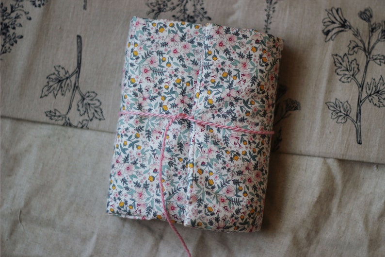 May include: A rectangular, floral-patterned fabric-covered item, wrapped with pink and white twine. The fabric features a white background with small pink, yellow, and green floral details. The item is resting on a beige fabric with a botanical print.