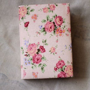 Puede incluir: Un libro con estampado floral, con rosas rosas, hojas verdes y peque&ntilde;as flores azules y amarillas sobre un fondo rosa claro. El libro rectangular est&aacute; cerrado.