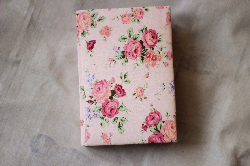 Puede incluir: Un libro con estampado floral, con rosas rosas, hojas verdes y peque&ntilde;as flores azules y amarillas sobre un fondo rosa claro. El libro rectangular est&aacute; cerrado.