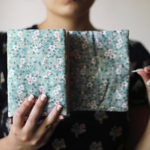 May include: A floral fabric journal with a teal background and white and pink flowers. The journal is open, revealing the inside pages. A white cord is held in the hand.
