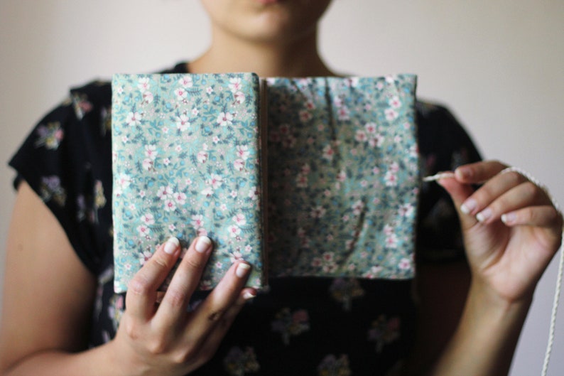 May include: A floral fabric journal with a teal background and white and pink flowers. The journal is open, revealing the inside pages. A white cord is held in the hand.
