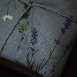 May include: A gray linen fabric item, possibly a sachet or small pillow, adorned with lavender and daisy floral patterns. The item is tied with a natural twine, suggesting a handmade or rustic aesthetic. The lavender blooms are purple, with green stems and leaves.