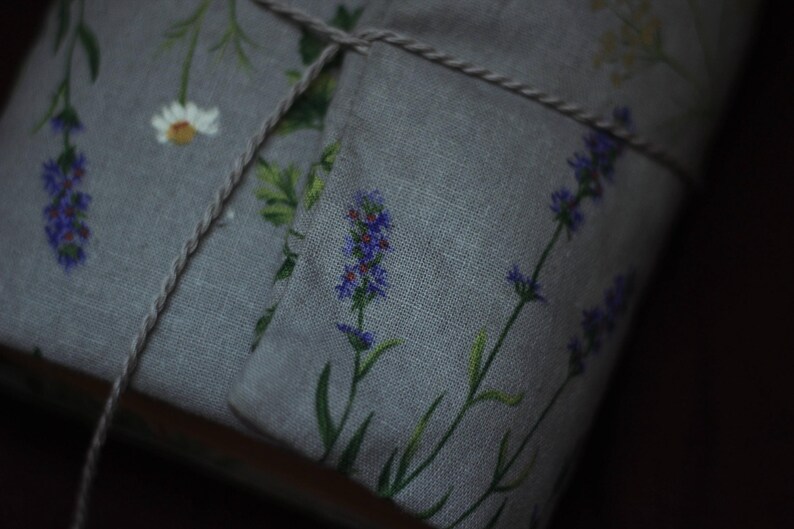 May include: A gray linen fabric item, possibly a sachet or small pillow, adorned with lavender and daisy floral patterns. The item is tied with a natural twine, suggesting a handmade or rustic aesthetic. The lavender blooms are purple, with green stems and leaves.