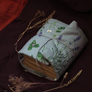 May include: A handmade book wrapped in a light blue fabric cover, adorned with illustrations of herbs and flowers. The cover is tied with a twine string. The book is resting on a dark brown surface, with dried sprigs nearby.