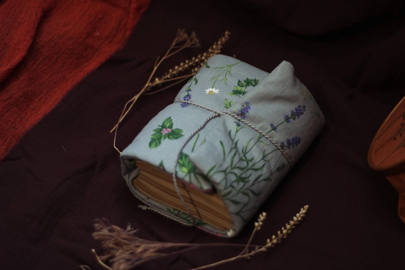 May include: A handmade book wrapped in a light blue fabric cover, adorned with illustrations of herbs and flowers. The cover is tied with a twine string. The book is resting on a dark brown surface, with dried sprigs nearby.
