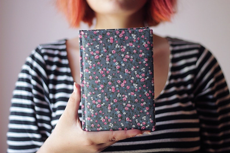 May include: A rectangular book with a floral pattern cover. The cover is a dark gray with a repeating pattern of pink roses, small white flowers, and blue leaves. The book is held in front of a person wearing a black and white striped shirt.