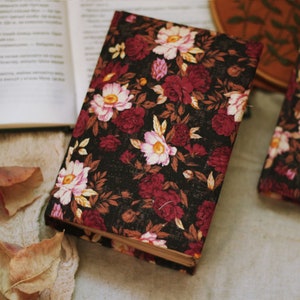 May include: A black fabric-covered journal with a floral pattern in shades of pink, red, and white. The journal is lying on a beige surface with brown leaves.