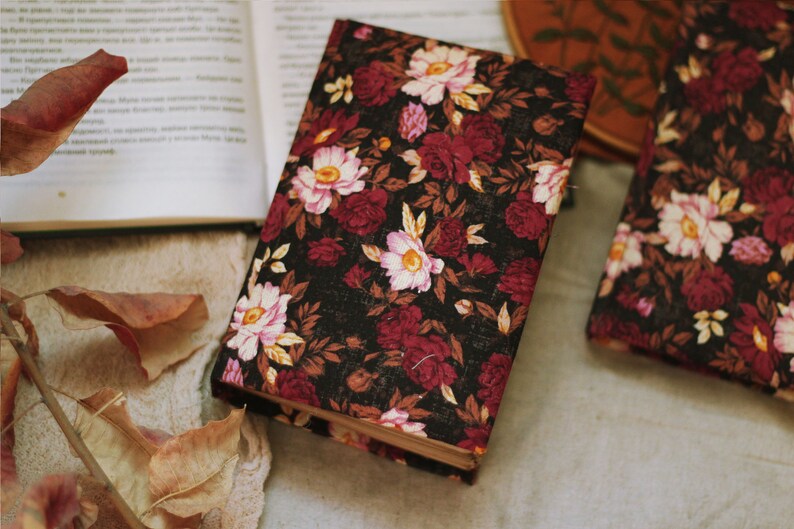 May include: A black fabric-covered journal with a floral pattern in shades of pink, red, and white. The journal is lying on a beige surface with brown leaves.