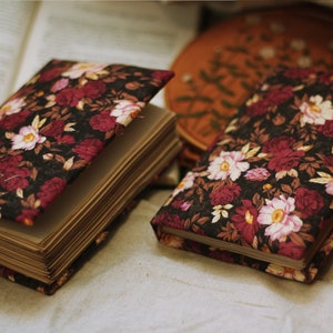 May include: Two closed journals with floral patterned covers. The covers are black with red and pink flowers. The journals are lying on a beige fabric surface.