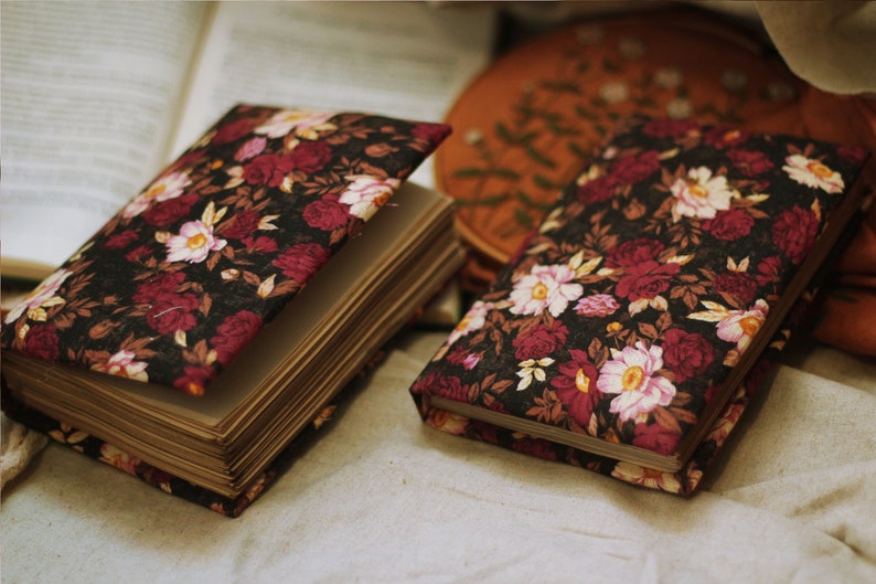 May include: Two closed journals with floral patterned covers. The covers are black with red and pink flowers. The journals are lying on a beige fabric surface.