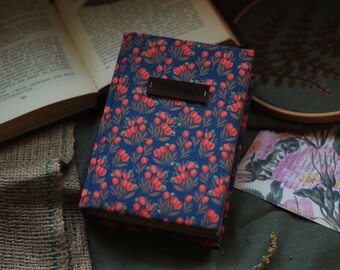 Puede incluir: Un cuaderno azul con un patrón floral y una etiqueta de cuero marrón que dice "Notebook". El cuaderno está sobre una superficie de arpillera.