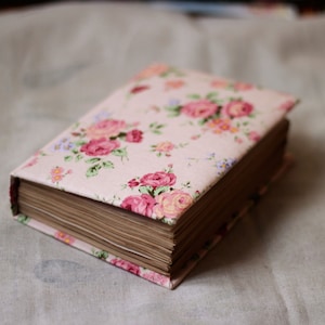 Puede incluir: Un libro cerrado con una cubierta estampada con flores. La cubierta es de color rosa claro con un estampado de rosas rosas y rojas, hojas verdes y peque&ntilde;as flores azules. Las p&aacute;ginas son de color marr&oacute;n claro.