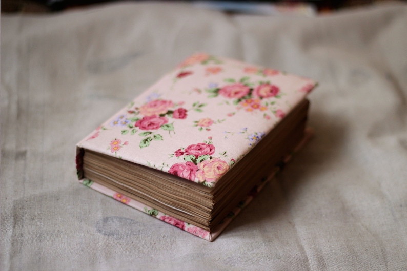 Puede incluir: Un libro cerrado con una cubierta estampada con flores. La cubierta es de color rosa claro con un estampado de rosas rosas y rojas, hojas verdes y peque&ntilde;as flores azules. Las p&aacute;ginas son de color marr&oacute;n claro.
