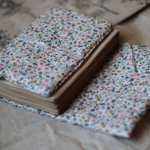 May include: A floral fabric-covered journal and matching book cover. The fabric features a white background with small pink, yellow, and blue flowers and green leaves. The journal is open, revealing aged paper pages.