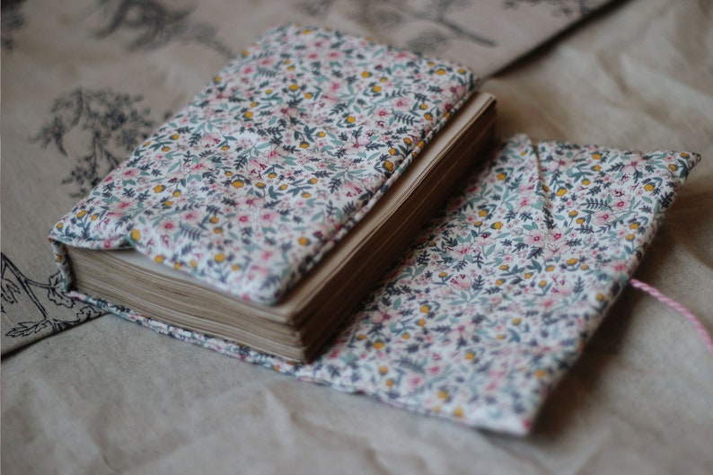 May include: A floral fabric-covered journal and matching book cover. The fabric features a white background with small pink, yellow, and blue flowers and green leaves. The journal is open, revealing aged paper pages.