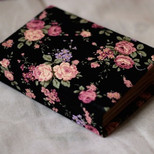 Puede incluir: Un libro rectangular con una cubierta floral negra con rosas rosas y moradas y hojas verdes. Las p&aacute;ginas del libro son visibles en el lateral y descansa sobre una superficie de tela blanca. El patr&oacute;n floral es detallado y vibrante.
