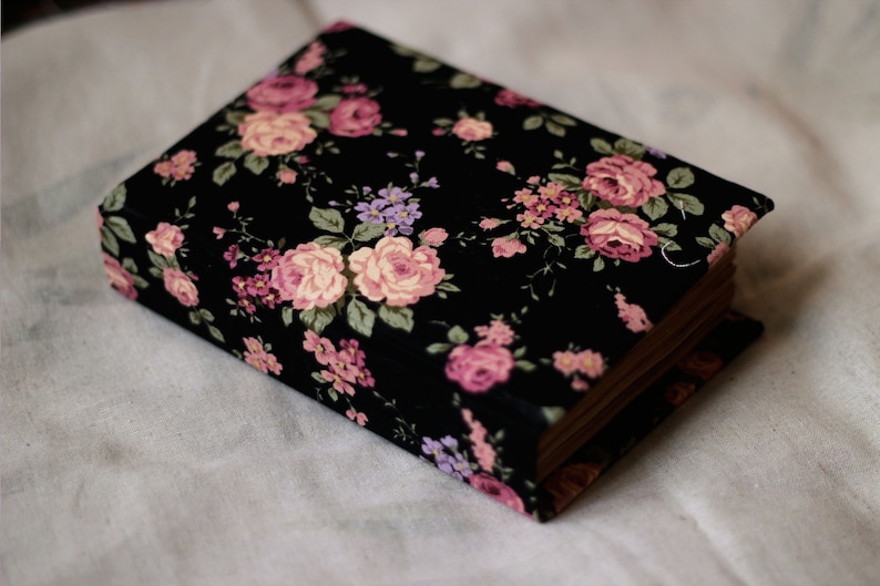 Puede incluir: Un libro rectangular con una cubierta floral negra con rosas rosas y moradas y hojas verdes. Las p&aacute;ginas del libro son visibles en el lateral y descansa sobre una superficie de tela blanca. El patr&oacute;n floral es detallado y vibrante.