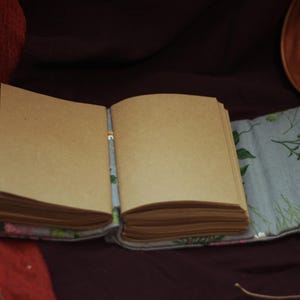 May include: An open, blank journal with tan-colored pages. The journal is resting on a light blue floral patterned fabric. A wooden decorative plate with floral embroidery is in the background.
