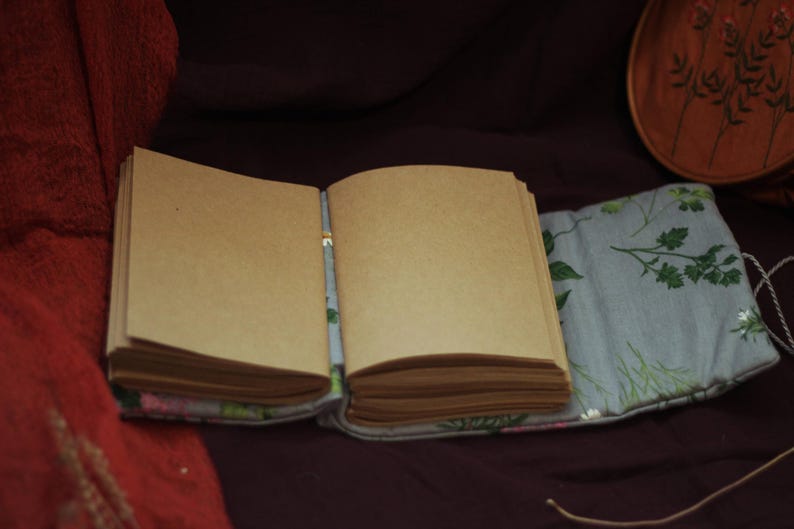 May include: An open, blank journal with tan-colored pages. The journal is resting on a light blue floral patterned fabric. A wooden decorative plate with floral embroidery is in the background.
