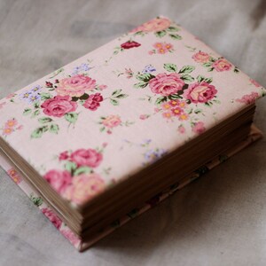 Puede incluir: Un libro cerrado con una cubierta de estampado floral. La cubierta es de color rosa claro con un patr&oacute;n repetido de rosas rosas y rojas, hojas verdes y peque&ntilde;as flores azules y amarillas. Las p&aacute;ginas son visibles y de color marr&oacute;n claro.