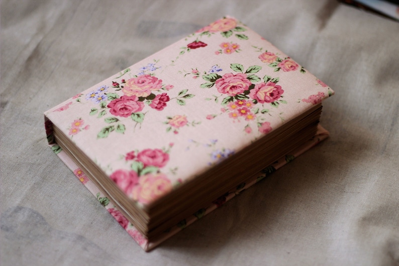 Puede incluir: Un libro cerrado con una cubierta de estampado floral. La cubierta es de color rosa claro con un patr&oacute;n repetido de rosas rosas y rojas, hojas verdes y peque&ntilde;as flores azules y amarillas. Las p&aacute;ginas son visibles y de color marr&oacute;n claro.