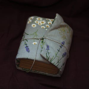 May include: A wrapped book with a floral fabric cover featuring daisies and lavender sprigs. The book is tied with twine and rests on a dark purple surface. The cover is light blue with white, green, and purple accents.