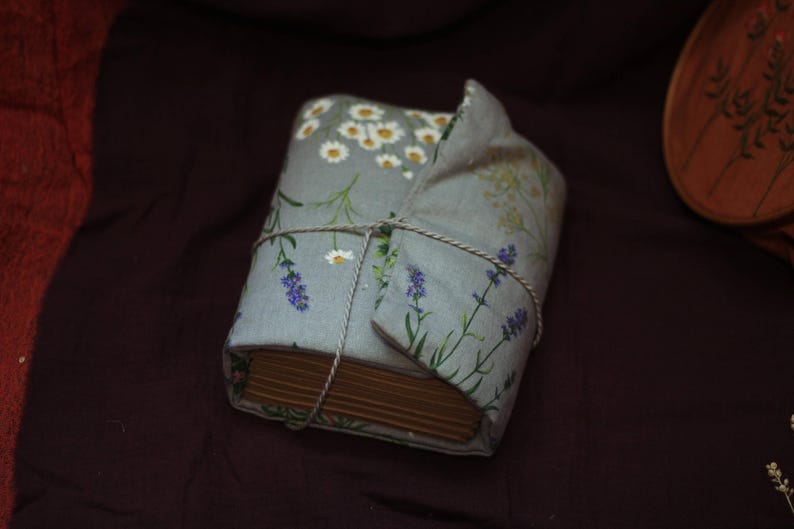 May include: A wrapped book with a floral fabric cover featuring daisies and lavender sprigs. The book is tied with twine and rests on a dark purple surface. The cover is light blue with white, green, and purple accents.
