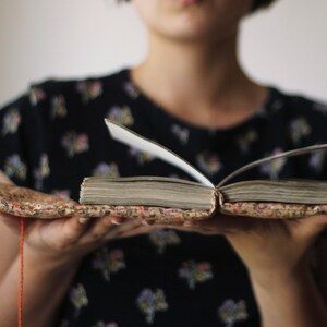 May include: An open book with aged, yellowed pages. The book is held open, revealing the text within. The cover is a patterned fabric with a floral design. An orange string is attached to the book.