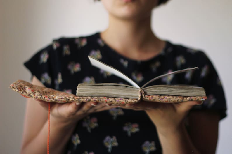 May include: An open book with aged, yellowed pages. The book is held open, revealing the text within. The cover is a patterned fabric with a floral design. An orange string is attached to the book.