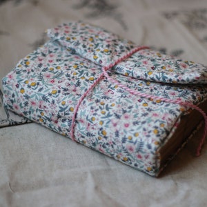 May include: A floral fabric-covered journal with a flap closure, tied with pink and white twine. The fabric features a pattern of small pink, yellow, and blue flowers on a white background. The journal is closed and resting on a neutral-toned surface.