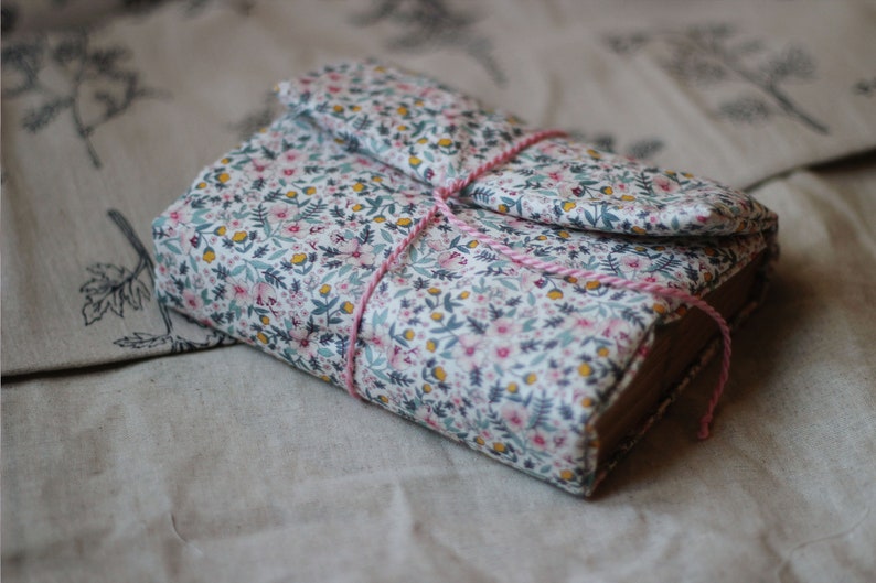 May include: A floral fabric-covered journal with a flap closure, tied with pink and white twine. The fabric features a pattern of small pink, yellow, and blue flowers on a white background. The journal is closed and resting on a neutral-toned surface.