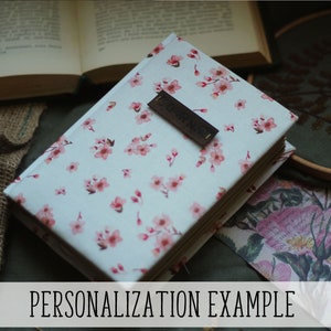 May include: A closed book with a floral pattern cover featuring pink blossoms on a white background. A dark brown rectangular tag is on the cover. The text "PERSONALIZATION EXAMPLE" is at the bottom of the image. Other books and decorative items are in the background.