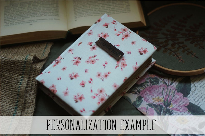 May include: A closed book with a floral pattern cover featuring pink blossoms on a white background. A dark brown rectangular tag is on the cover. The text "PERSONALIZATION EXAMPLE" is at the bottom of the image. Other books and decorative items are in the background.