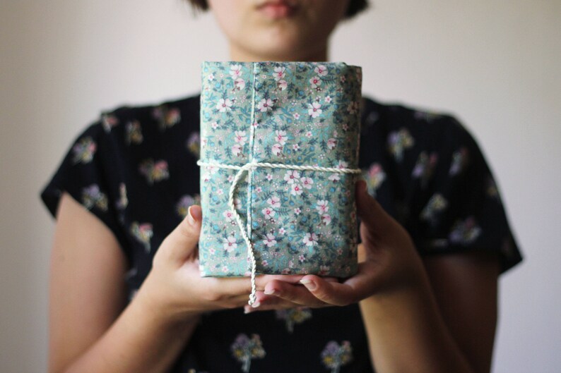 May include: A rectangular package wrapped in teal fabric with a floral pattern of white and pink flowers. The package is tied with a white string. The person holding the package is wearing a black floral top.
