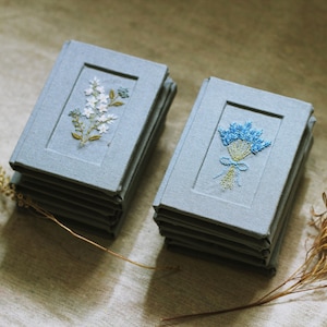 May include: Two stacks of small, light blue fabric-covered notebooks. The left stack features an embroidered white floral sprig with green leaves. The right stack displays an embroidered blue flower bouquet tied with a light blue ribbon.