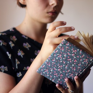 May include: A floral-patterned journal with a blue cover and pink and white flowers. The pages are aged, and the journal is being held open. The person is wearing a dark blue top with a floral pattern.