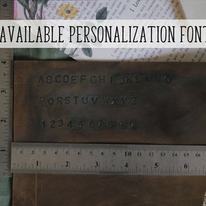 May include: A brown leather rectangle displays stamped alphabet letters and numbers, with a ruler showing inches. The text "AVAILABLE PERSONALIZATION FONT" is at the top. The image suggests customization options for leather goods.