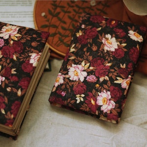 May include: Two floral patterned journals with burgundy and white flowers on a black background. The journals are closed and lying on a white fabric surface.