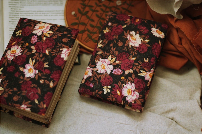 May include: Two floral patterned journals with burgundy and white flowers on a black background. The journals are closed and lying on a white fabric surface.