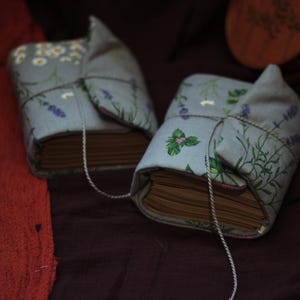 May include: Two fabric-covered books with floral and herb patterns. The covers are light blue with illustrations of white flowers, lavender, and green herbs. Each book is tied with a string. The books are on a dark purple surface.