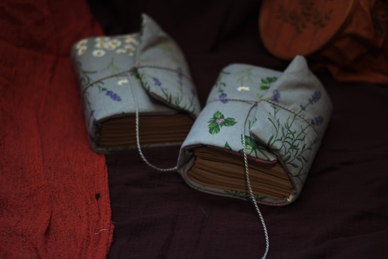 May include: Two fabric-covered books with floral and herb patterns. The covers are light blue with illustrations of white flowers, lavender, and green herbs. Each book is tied with a string. The books are on a dark purple surface.