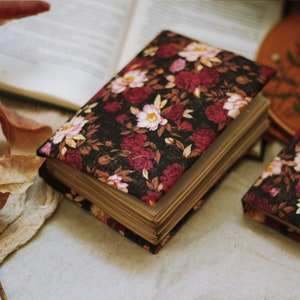 May include: Two closed hardback journals with floral patterned covers. The covers are black with red and pink flowers. The journals are lying on a beige surface.