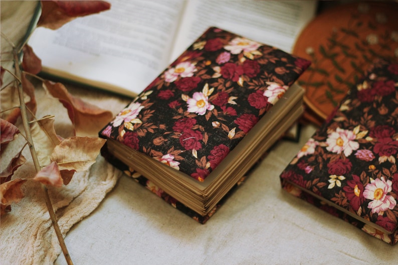 May include: Two closed hardback journals with floral patterned covers. The covers are black with red and pink flowers. The journals are lying on a beige surface.