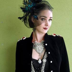May include: A woman wearing a black velvet jacket with silver and black beaded necklaces. She is wearing a black veil with blue and green feathers on top of her head.